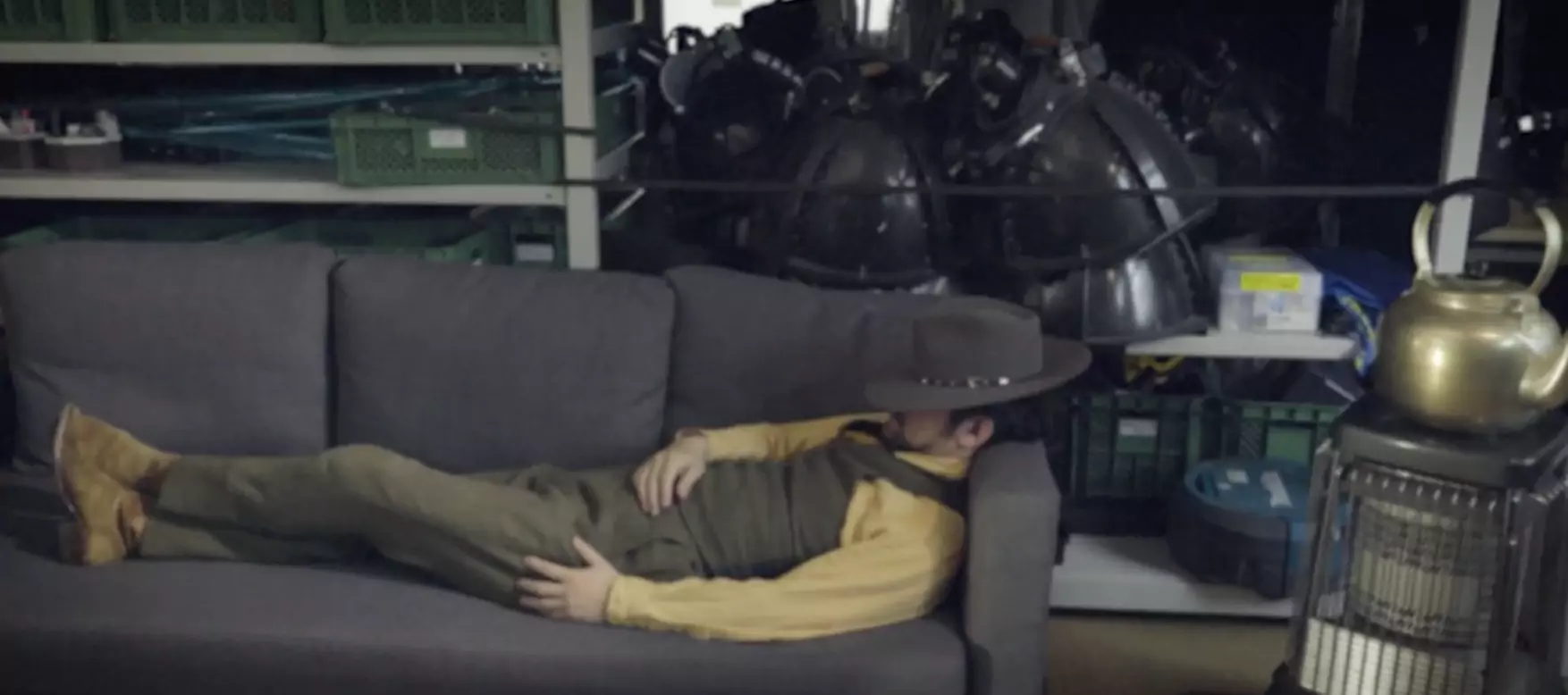 Screen capture from The Memphis Bell’s music video, showing a man in a stylish suit with cowboy had and boots lying on a couch in a warehouse filled with large lighting fixtures and a space heater with a large kettle. 